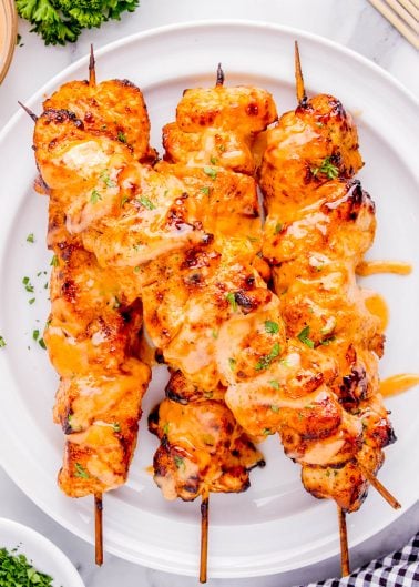 Three grilled chicken skewers drizzled with orange sauce and garnished with chopped herbs, served on a white plate.