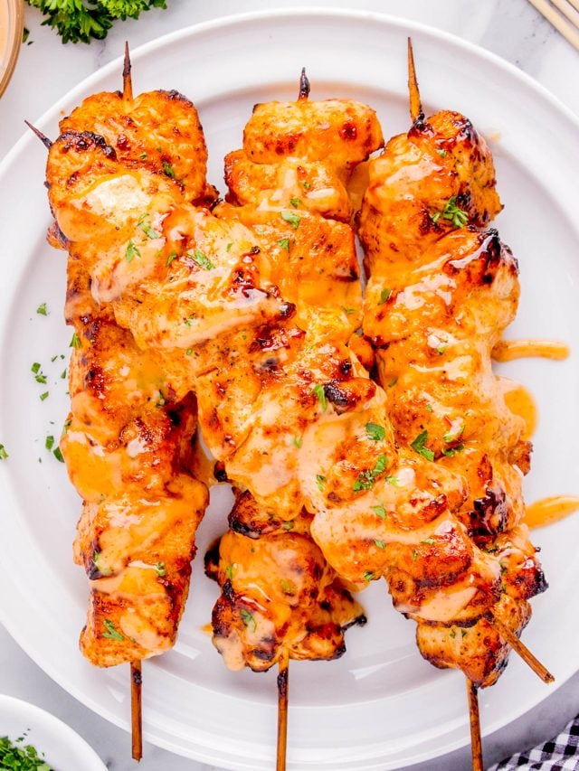Three grilled chicken skewers drizzled with orange sauce and garnished with chopped herbs, served on a white plate.