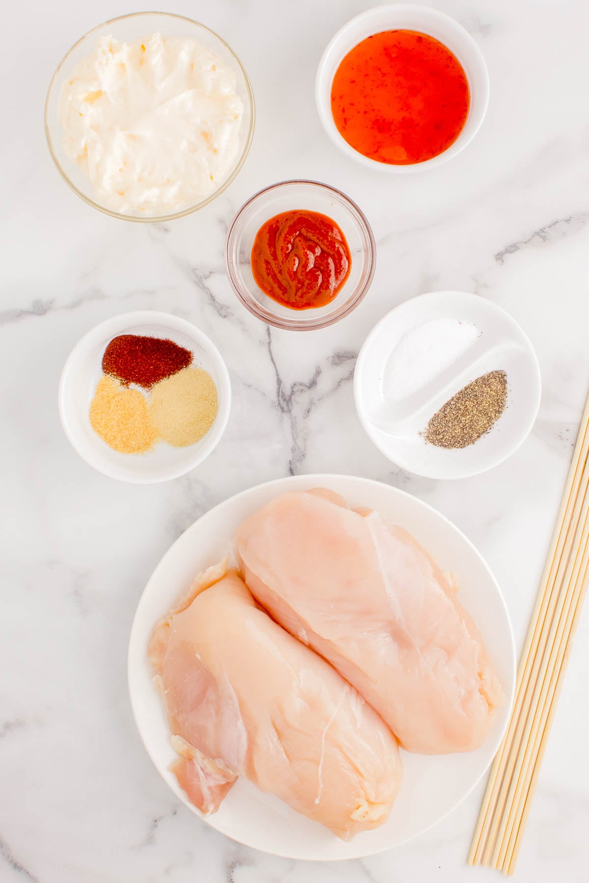 Raw chicken breasts on a plate surrounded by small bowls containing mayonnaise, sweet chili sauce, ketchup, spices, salt, and pepper, all on a marble surface with wooden skewers nearby.