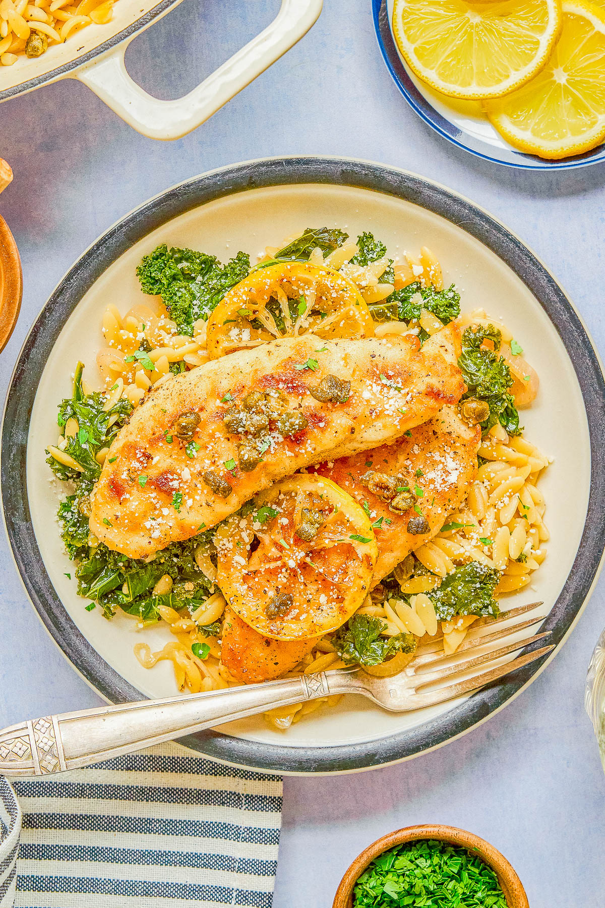 A plate with breaded chicken breast, lemon slices, orzo pasta, kale, and capers, garnished with grated cheese and herbs, next to a fork and a bowl of lemon slices.