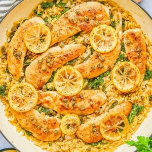 Cooked chicken breasts topped with lemon slices and capers, served over orzo pasta with spinach in a large white skillet. A small bowl of grated cheese and lemon wedges are nearby.