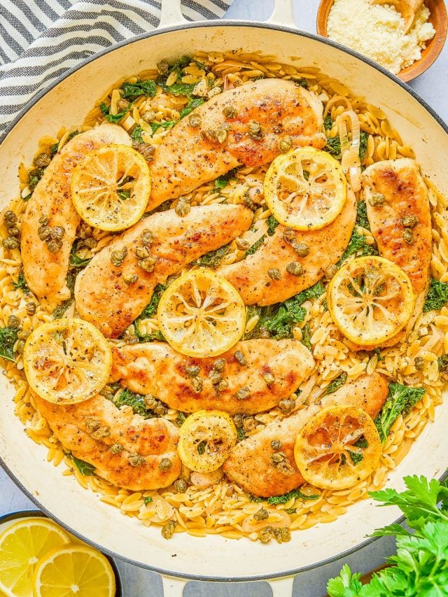 Cooked chicken breasts topped with lemon slices and capers, served over orzo pasta with spinach in a large white skillet. A small bowl of grated cheese and lemon wedges are nearby.