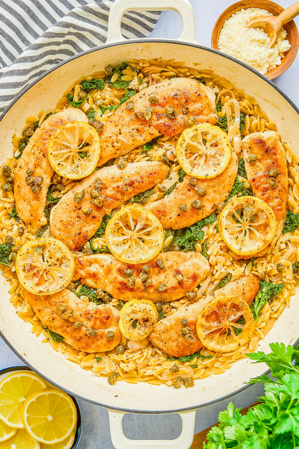 Cooked chicken breasts topped with lemon slices and capers, served over orzo pasta with spinach in a large white skillet. A small bowl of grated cheese and lemon wedges are nearby.