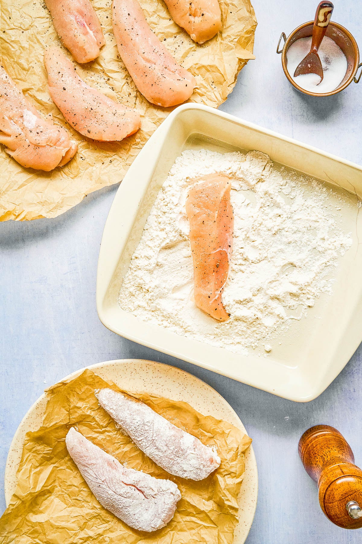 Raw chicken tenders being coated in flour; seasoned tenders on parchment, a dish with flour, and prepped coated pieces on a plate. Salt and pepper are nearby.