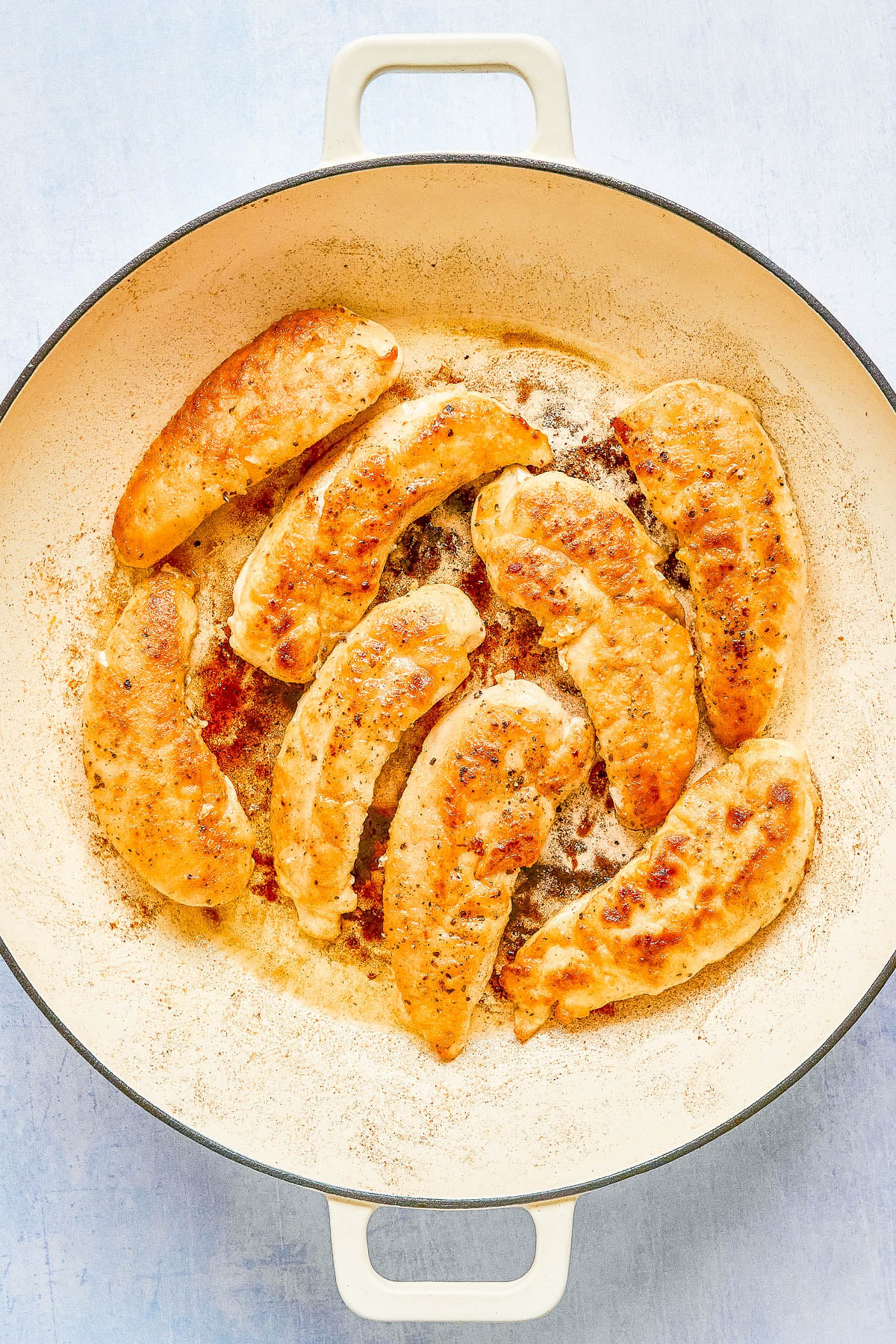 Cooked chicken tenders are arranged in a single layer inside a large, round cream-colored skillet on a light surface.