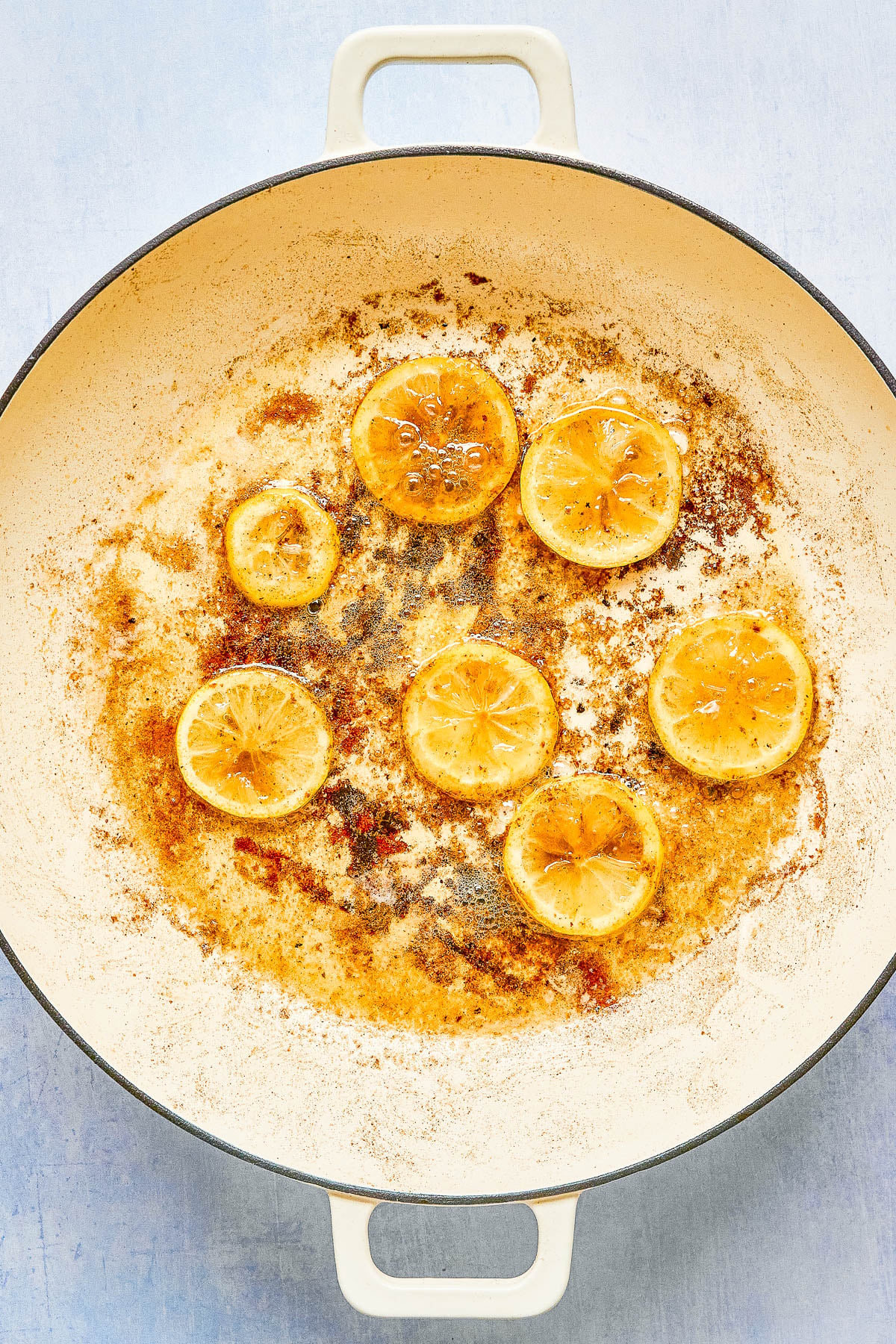 Sliced lemons are seared and caramelized in a large white enamel pan with browned residue and oily streaks on the surface.