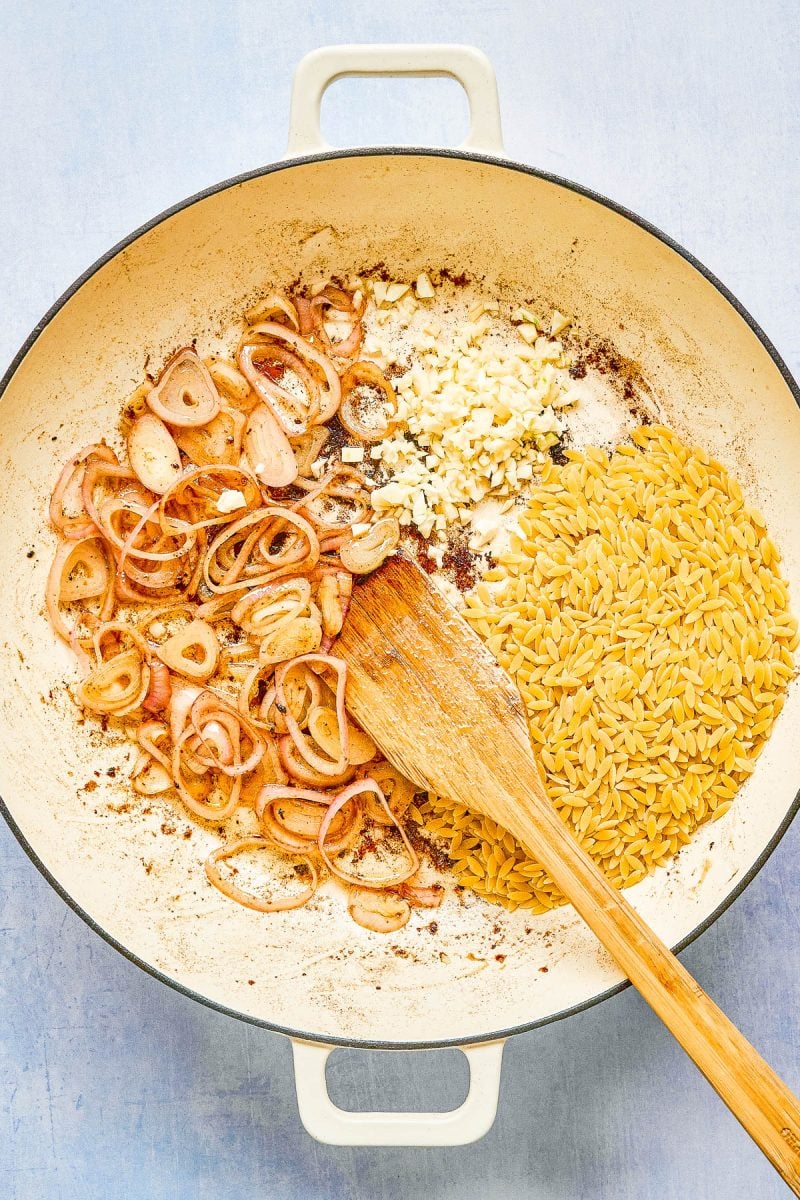 A large white pan containing sliced shallots, minced garlic, and uncooked orzo with a wooden spatula stirring the ingredients.