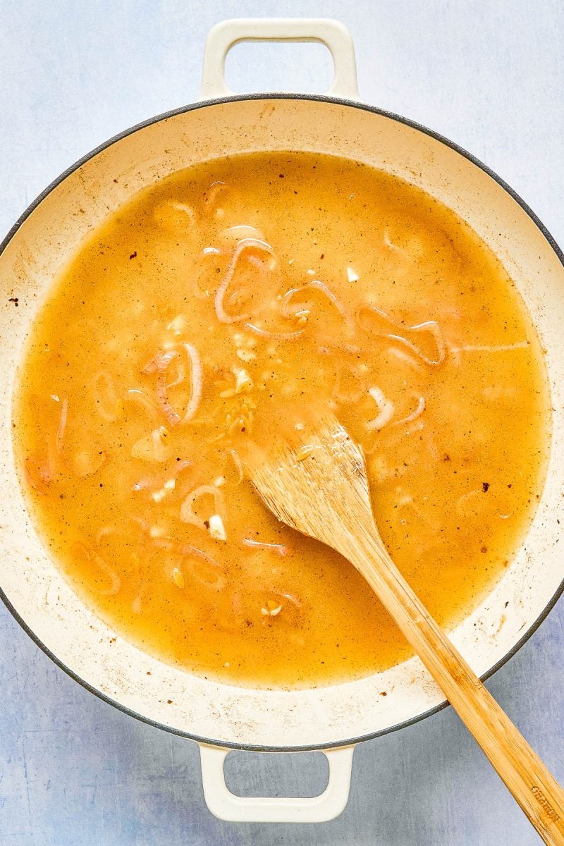 A light brown broth with sliced onions simmers in a white pot, stirred by a wooden spoon on a light blue surface.
