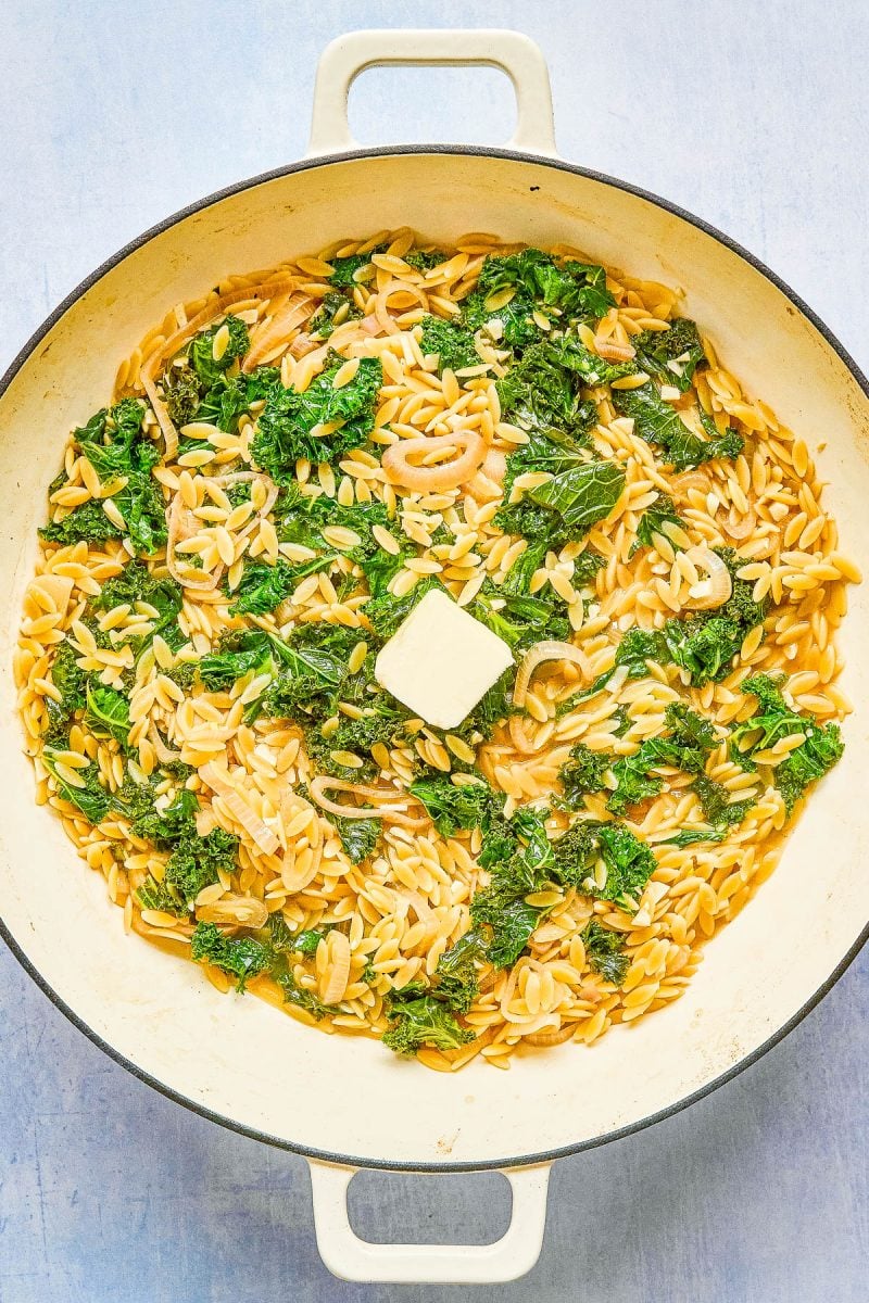 A large white skillet filled with cooked orzo pasta, wilted greens, onions, and a pat of butter in the center.