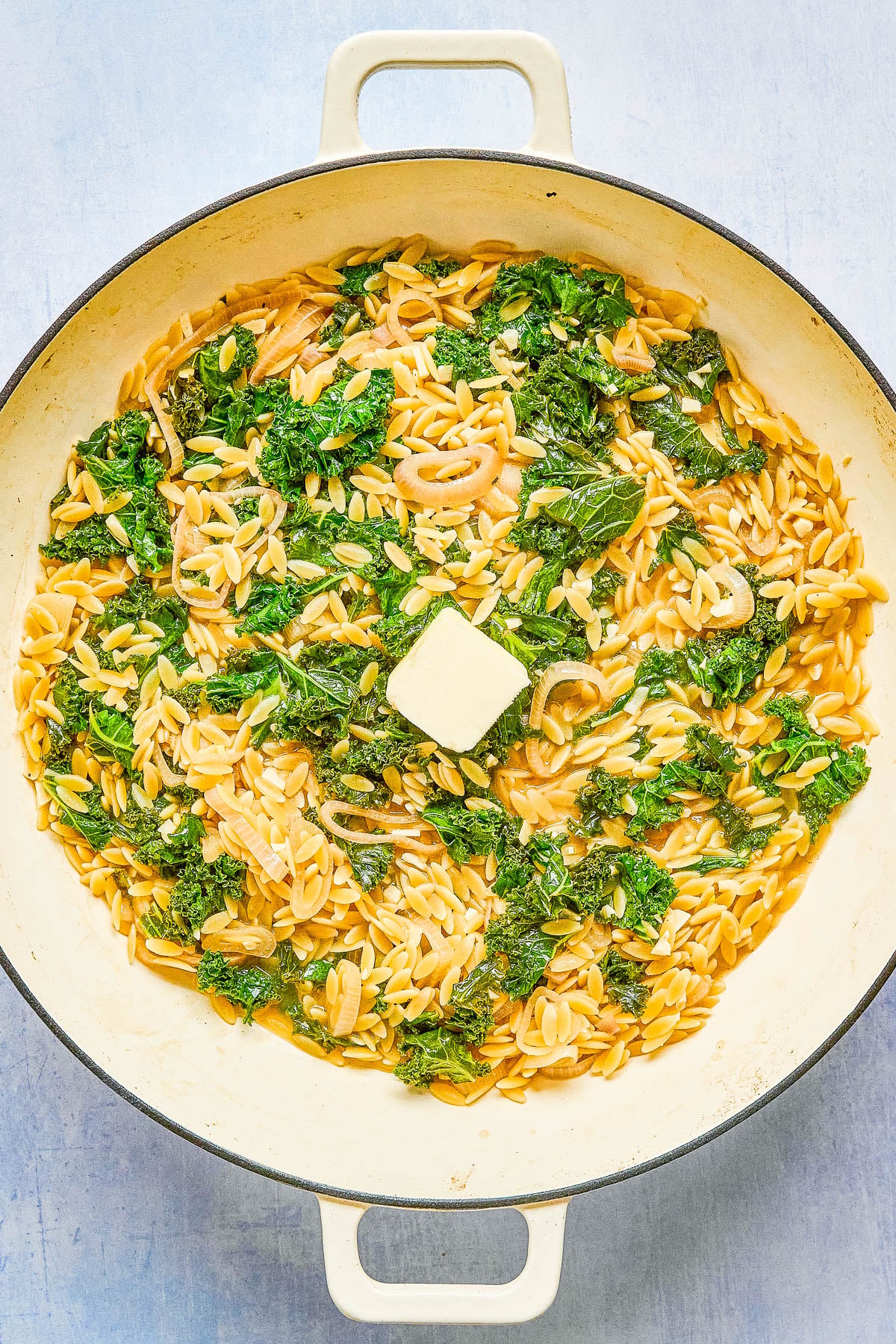 A large white skillet filled with cooked orzo pasta, wilted greens, onions, and a pat of butter in the center.
