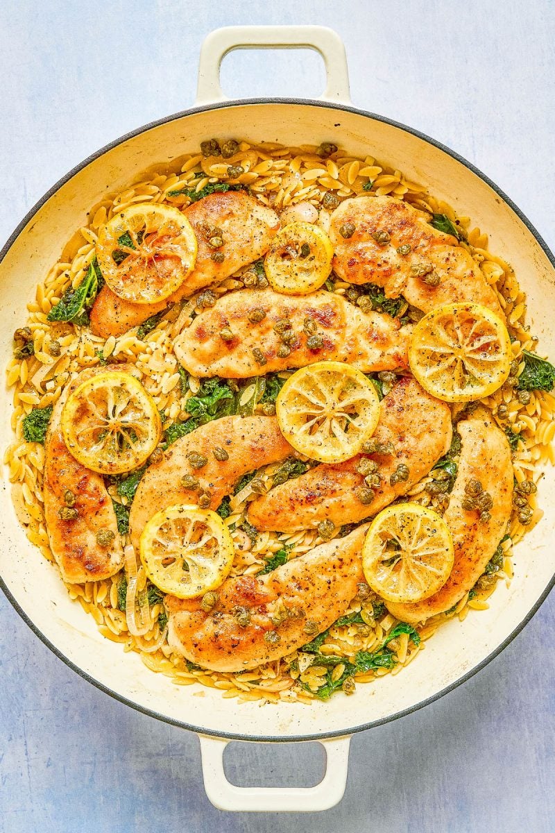 A large pan filled with cooked chicken breasts, orzo pasta, spinach, capers, and lemon slices, arranged neatly on a light surface.