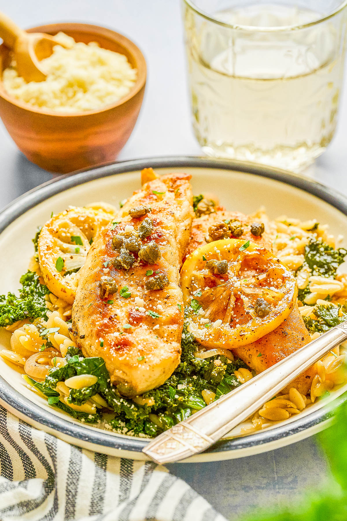 A plate of cooked chicken breast with lemon slices, served on a bed of orzo pasta and kale, with a fork, a glass of white wine, and a bowl of grated cheese in the background.