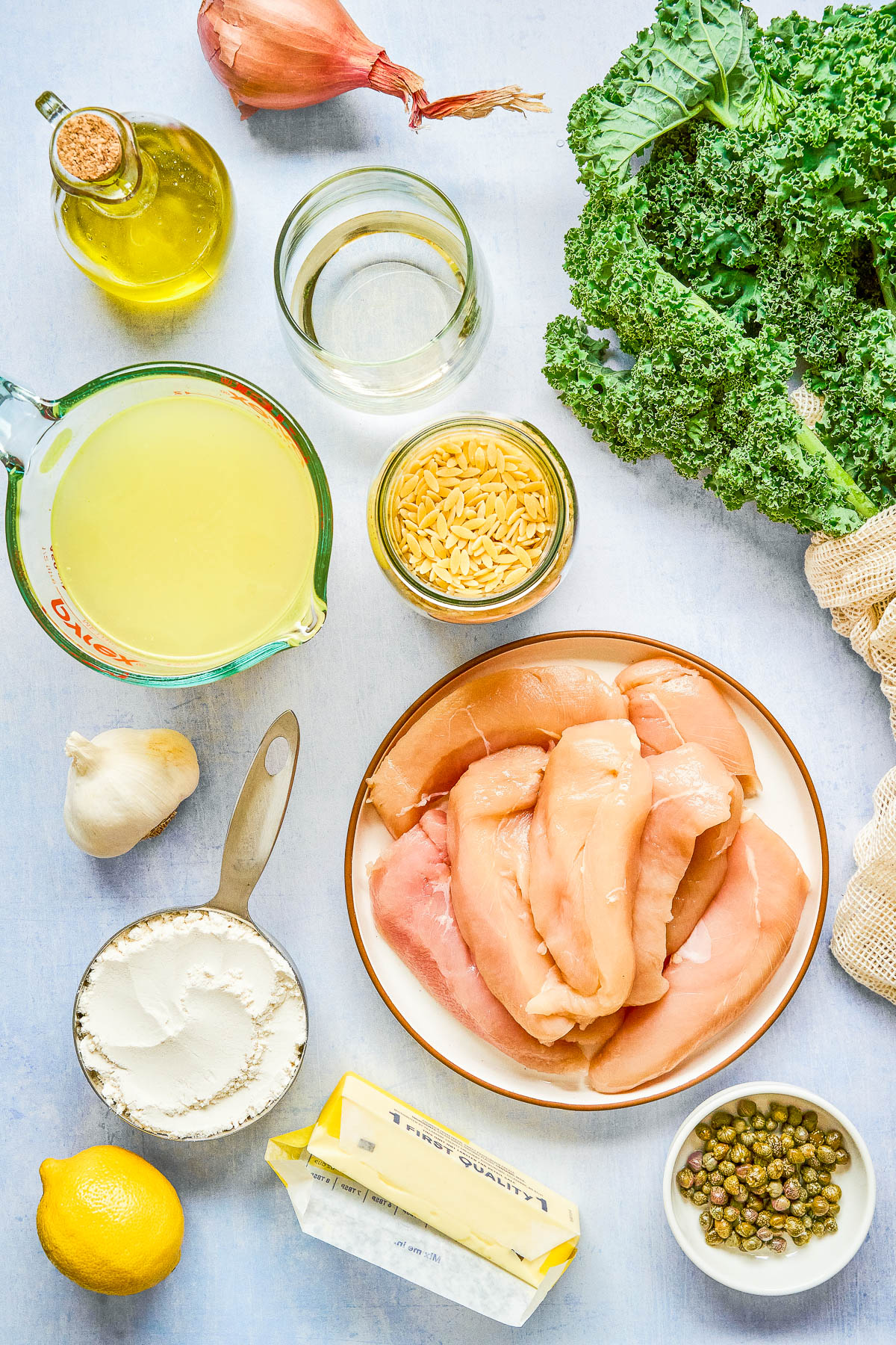 Raw chicken breasts, kale, orzo, garlic, shallot, lemon, capers, flour, butter, olive oil, chicken broth, and white wine arranged on a light surface.