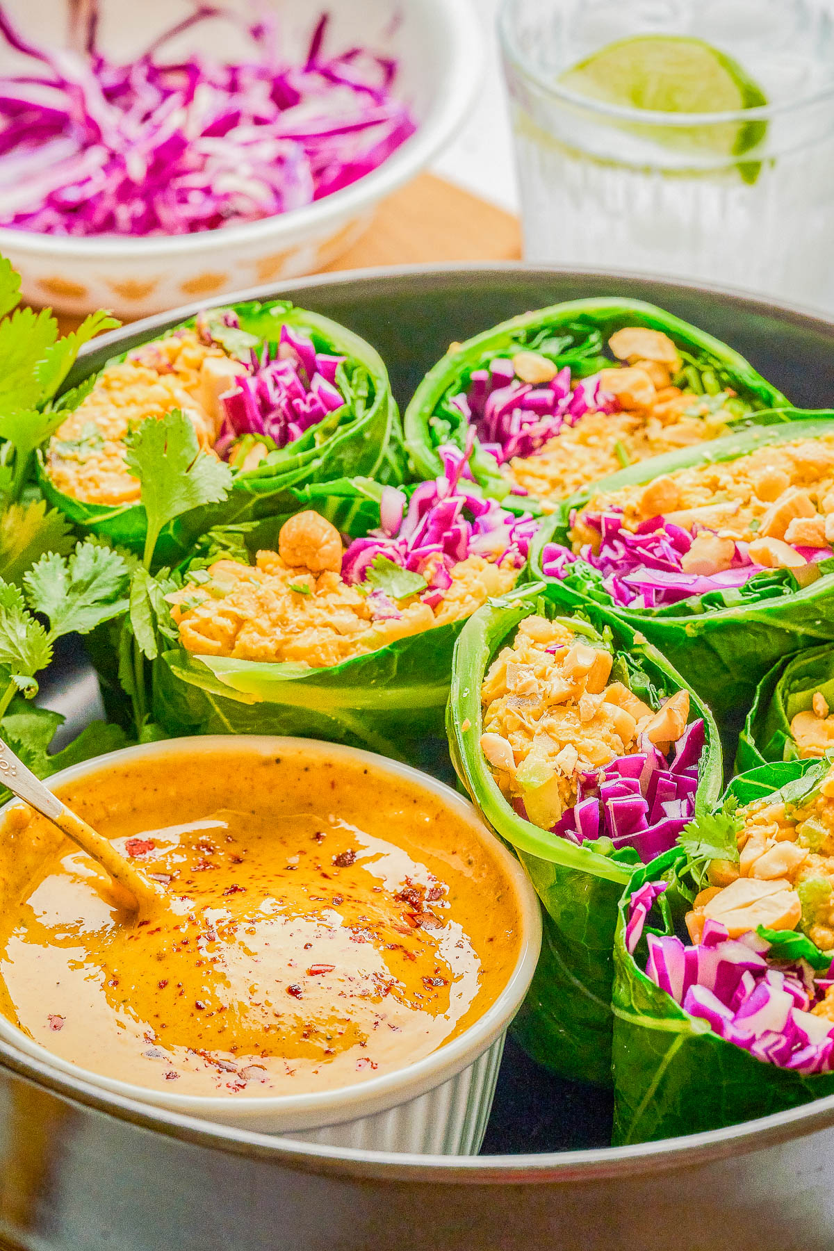 Fresh vegetable wraps with shredded purple cabbage and peanuts, served with a bowl of dipping sauce and a glass of water with lime in the background.