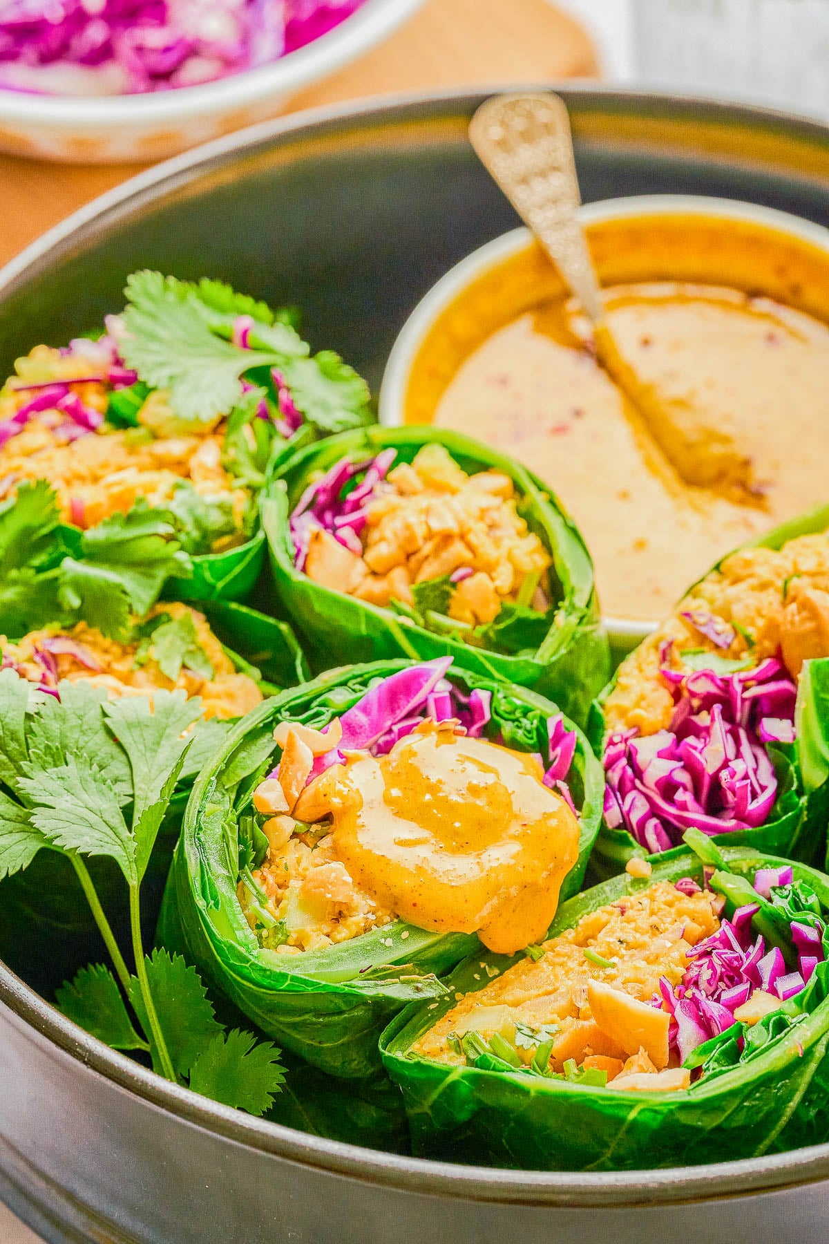Collard green wraps filled with vegetables and rice, garnished with cilantro and purple cabbage, served with a bowl of creamy yellow dipping sauce.