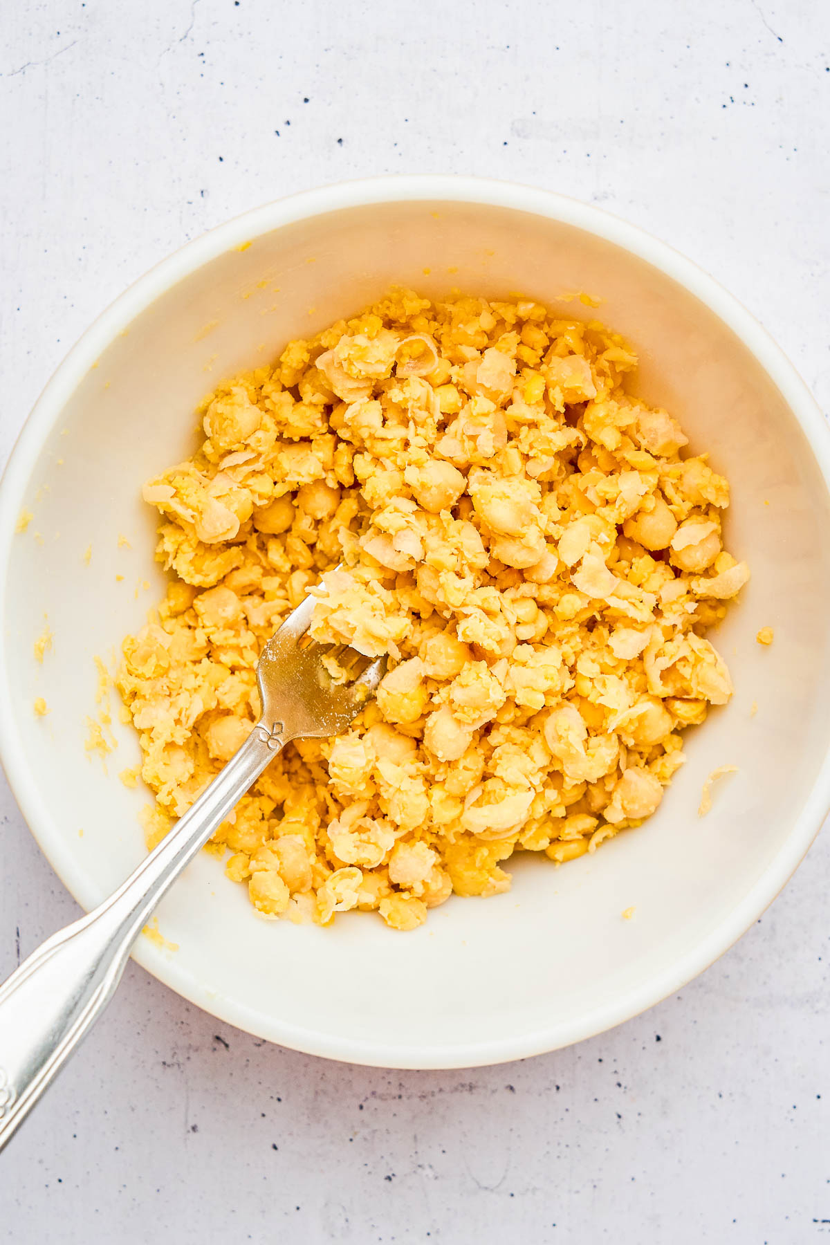 A white bowl containing mashed chickpeas with a fork resting inside, on a light speckled surface.