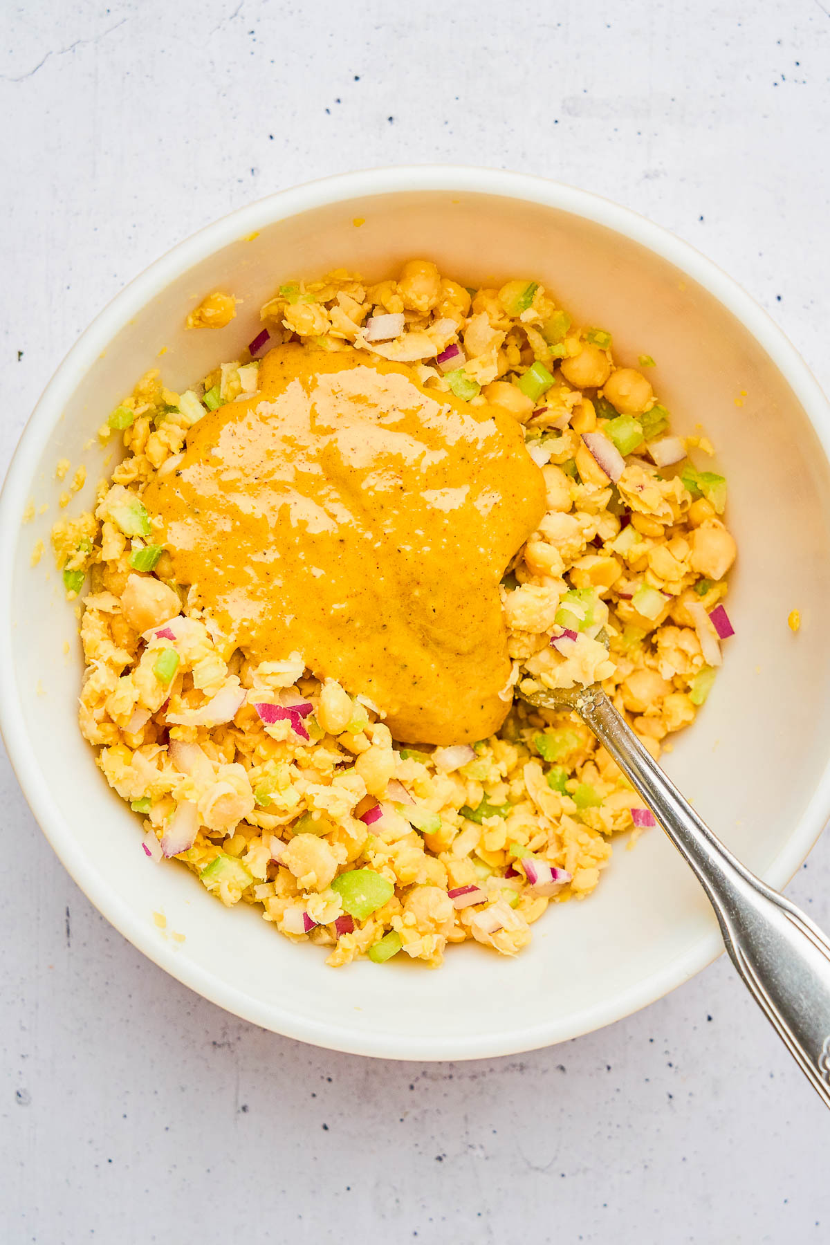 A white bowl containing a mixture of chickpeas, chopped celery, and red onion, topped with a yellow sauce, with a metal spoon resting inside.