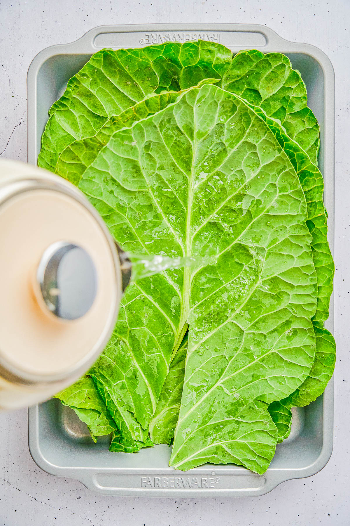 A kettle pours hot water over large green cabbage leaves arranged in a metal baking pan.