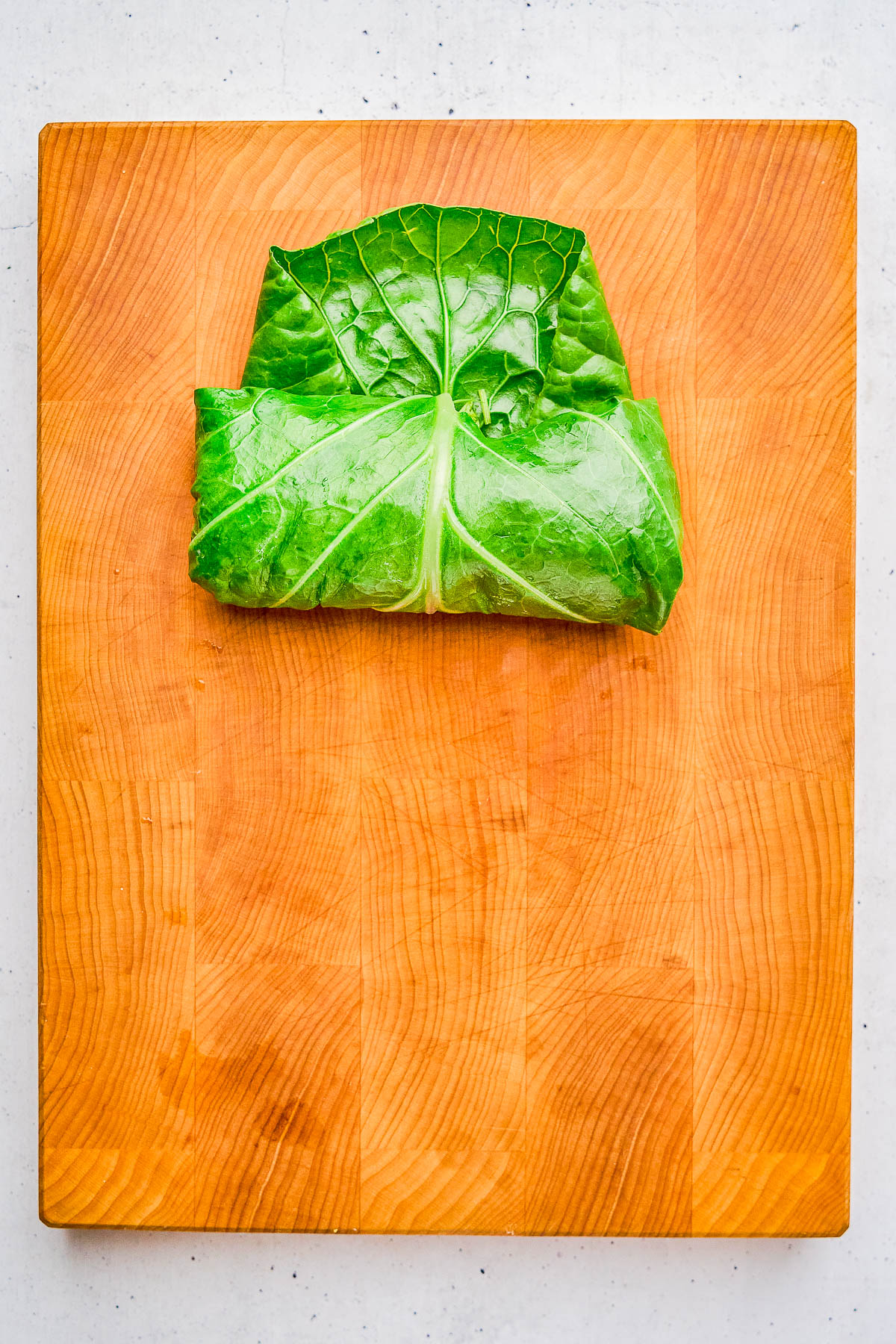 A large green leaf is wrapped around a rectangular filling and placed on a wooden cutting board.