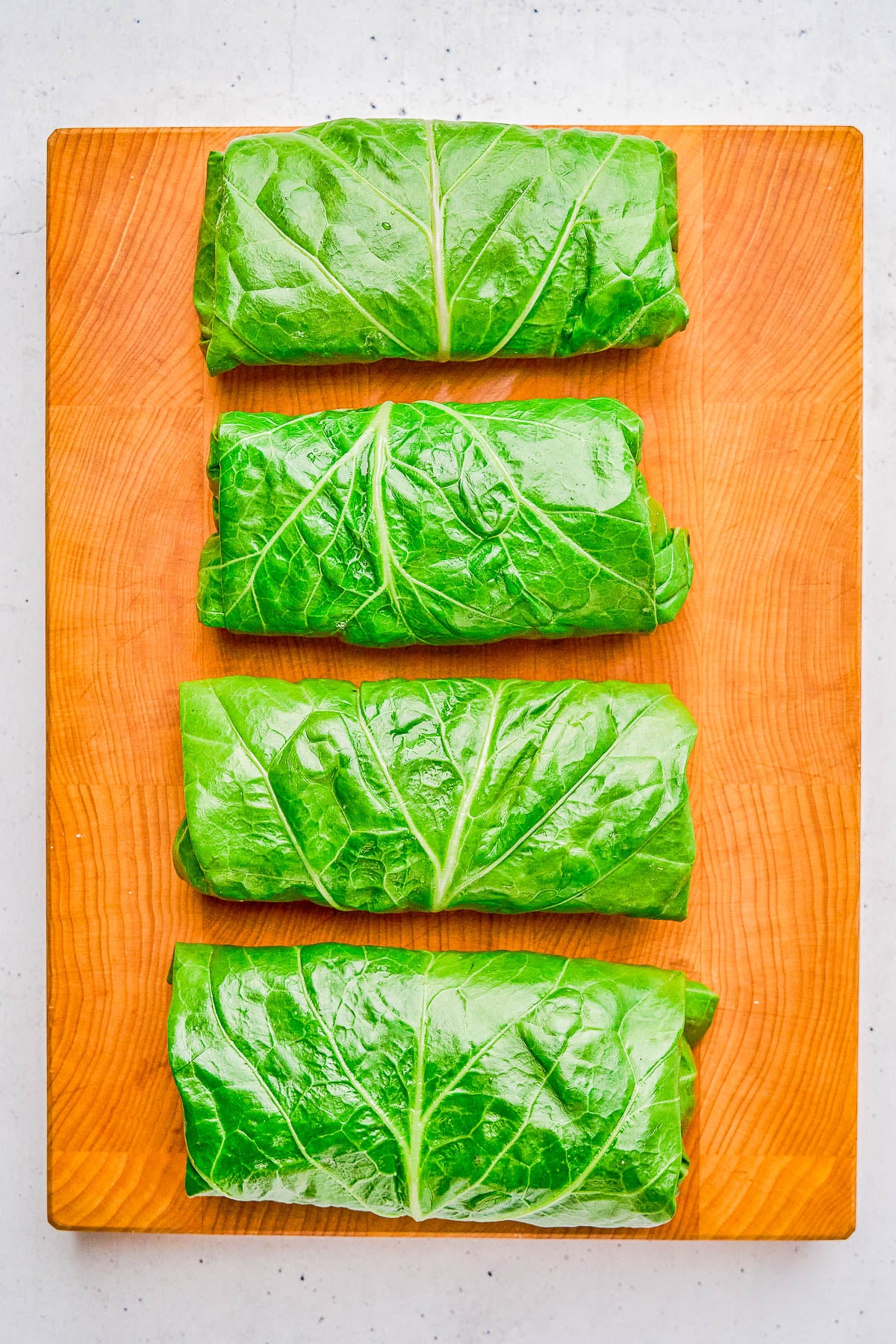 Four collard green wraps are neatly arranged in a vertical row on a wooden cutting board.