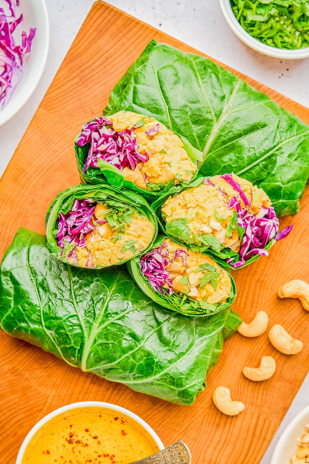 Collard green wraps filled with chickpeas, shredded purple cabbage, leafy greens, and herbs on a wooden board, with cashews and a bowl of yellow dipping sauce nearby.