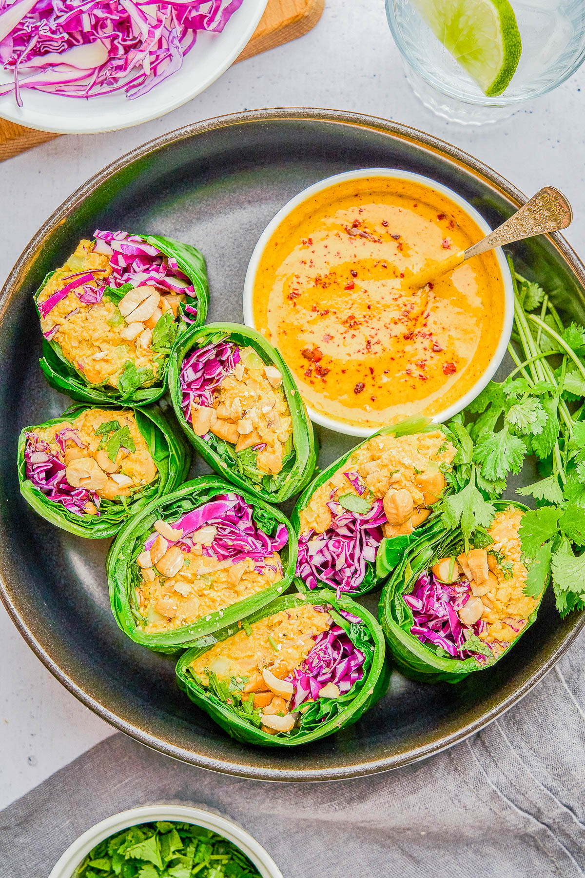 A plate of fresh lettuce wraps filled with shredded purple cabbage and nuts is served with a bowl of creamy orange dipping sauce, garnished with herbs.