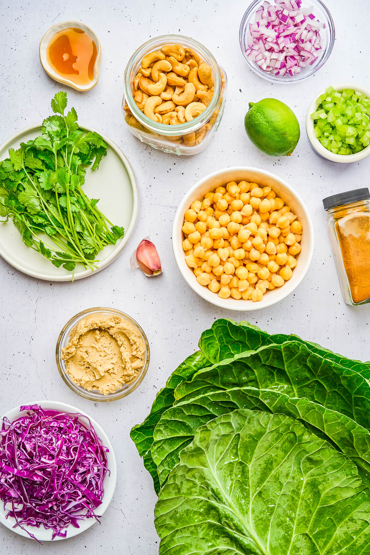 Assorted fresh ingredients including chickpeas, cashews, cilantro, red cabbage, celery, red onion, lime, hummus, a garlic clove, honey, and collard green leaves arranged on a white surface.