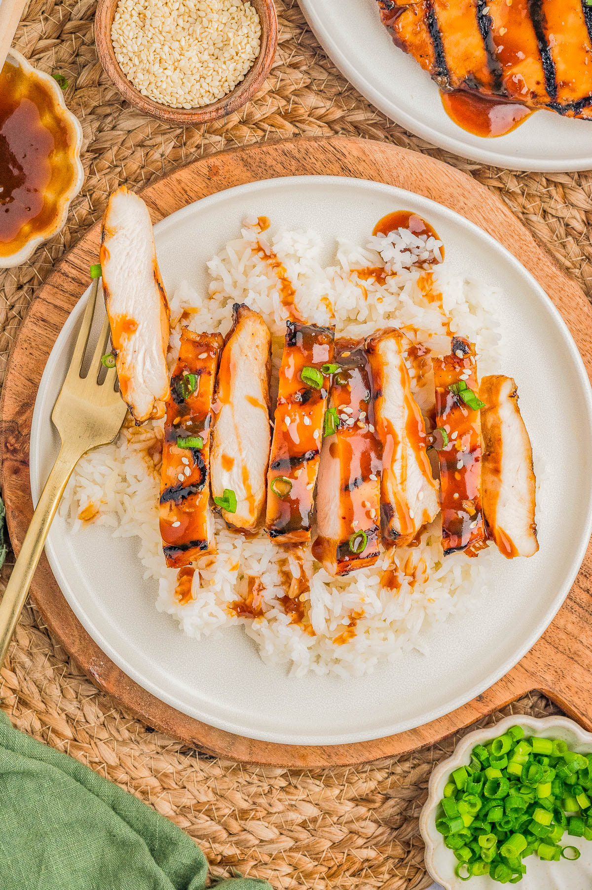 A plate of sliced grilled chicken with sauce, served over white rice and garnished with chopped green onions. A fork is placed on the side of the plate.