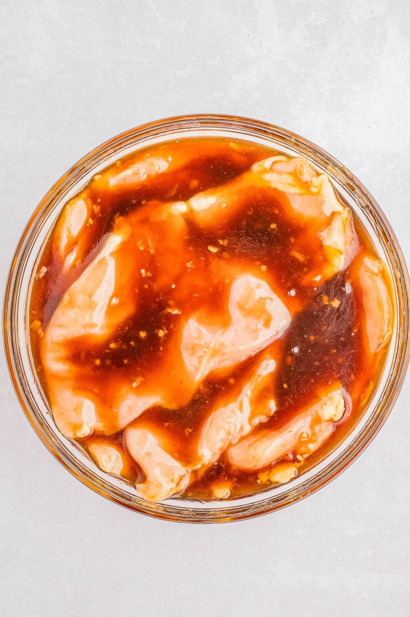 Raw chicken pieces marinating in a reddish-brown sauce inside a glass bowl, viewed from above on a light surface.