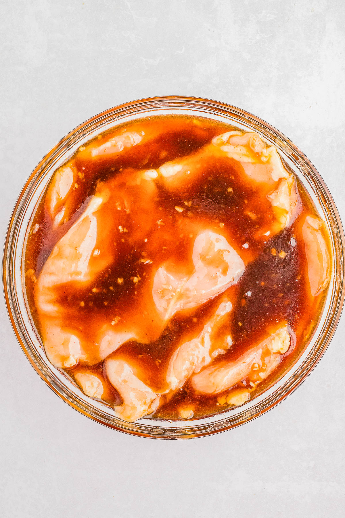 Raw chicken pieces marinating in a reddish-brown sauce inside a glass bowl, viewed from above on a light surface.