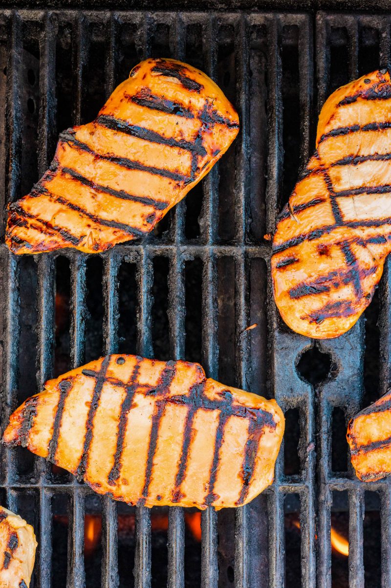 Four pieces of grilled chicken breast with visible char marks cooking on a barbecue grill.