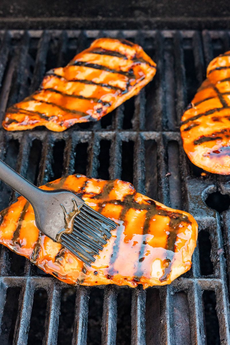 Three pieces of chicken are being grilled, with one piece being brushed with barbecue sauce using a silicone basting brush.