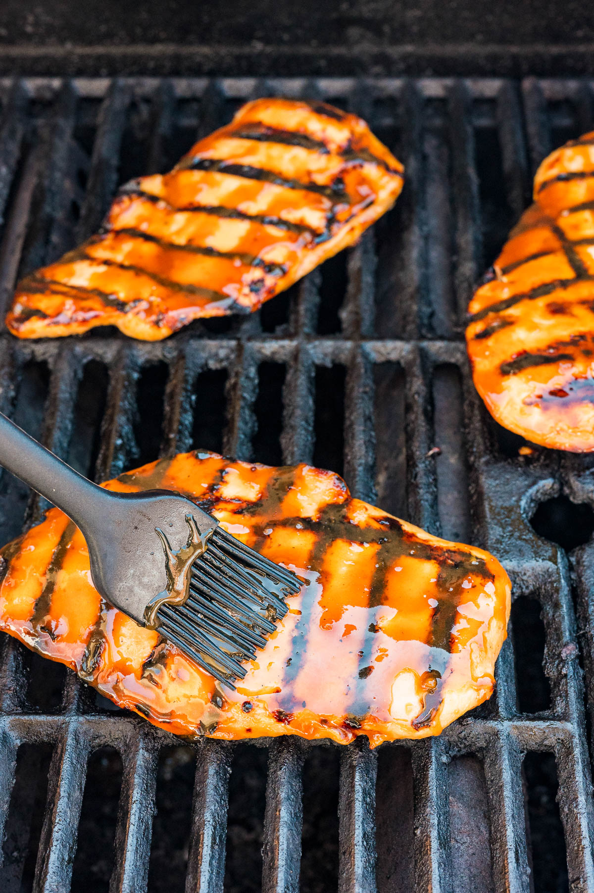 Three pieces of chicken are being grilled, with one piece being brushed with barbecue sauce using a silicone basting brush.