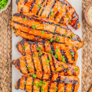 Four grilled chicken breasts with a glossy glaze, topped with chopped green onions and sesame seeds, are arranged on a rectangular white platter.