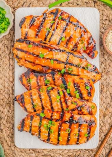 Four grilled chicken breasts with a glossy glaze, topped with chopped green onions and sesame seeds, are arranged on a rectangular white platter.