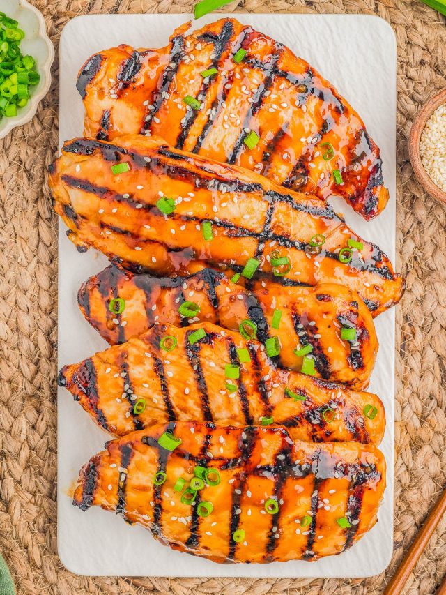 Four grilled chicken breasts with a glossy glaze, topped with chopped green onions and sesame seeds, are arranged on a rectangular white platter.