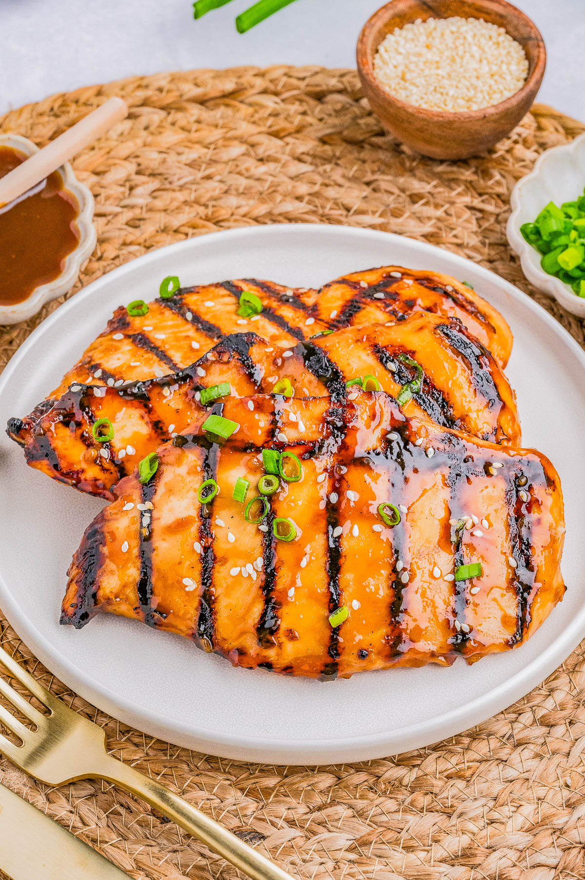 Two glazed, grilled chicken breasts garnished with sesame seeds and chopped green onions on a white plate, with condiments and utensils surrounding them.