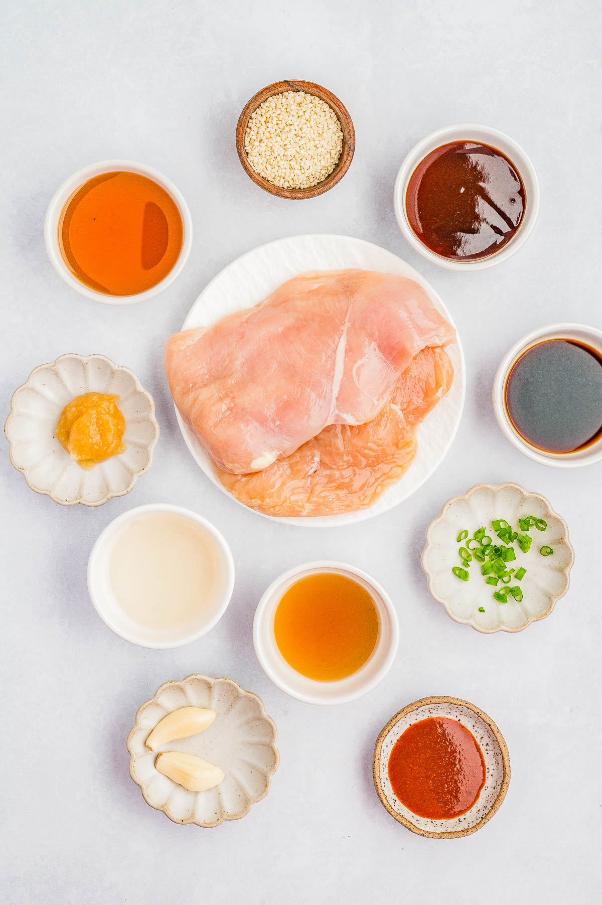 Raw chicken breast on a plate surrounded by bowls of various sauces, seasonings, sliced green onions, sesame seeds, and garlic cloves on a white surface.