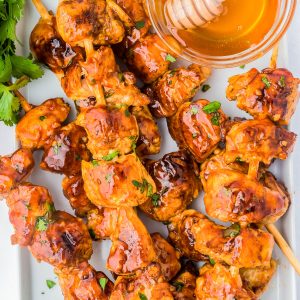 Grilled chicken skewers glazed with sauce, served with a bowl of honey and a honey dipper, garnished with fresh cilantro and a slice of lime.