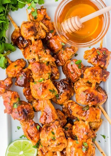 Grilled chicken skewers glazed with sauce, served with a bowl of honey and a honey dipper, garnished with fresh cilantro and a slice of lime.