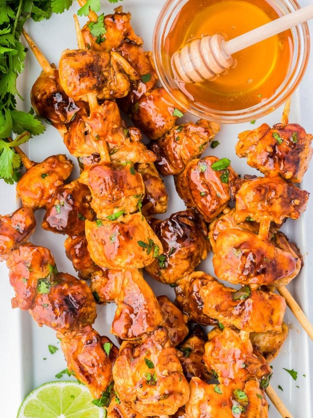 Grilled chicken skewers glazed with sauce, served with a bowl of honey and a honey dipper, garnished with fresh cilantro and a slice of lime.