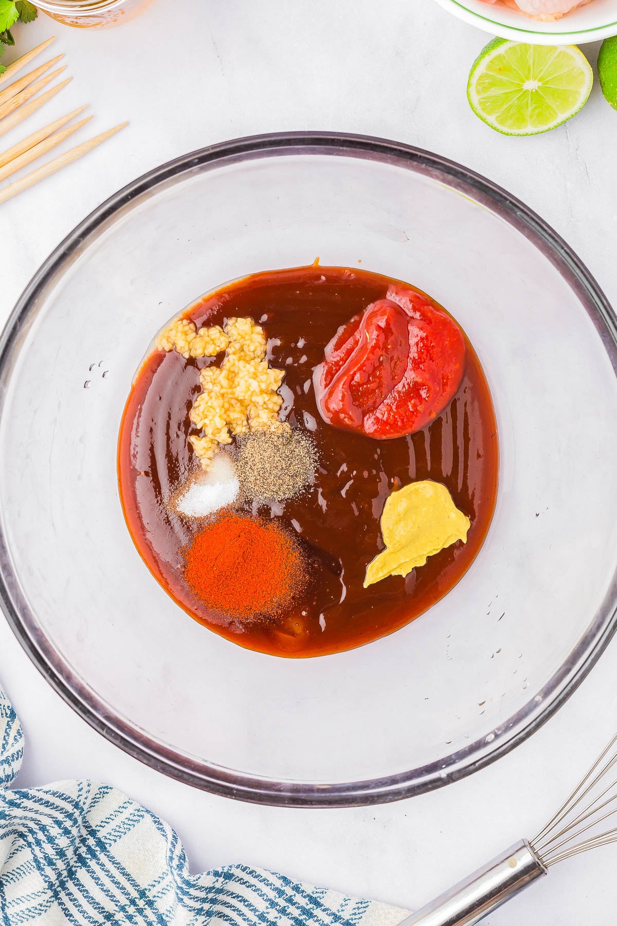 A clear bowl containing brown sauce, minced garlic, ketchup, yellow mustard, salt, pepper, and paprika, with a whisk and a blue plaid cloth nearby.