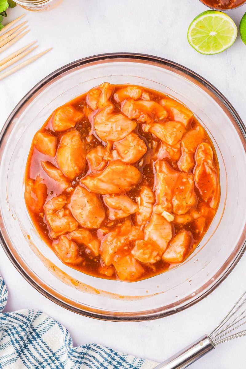 Raw chicken pieces marinating in a reddish sauce in a glass bowl, with a whisk, skewers, lime halves, and a blue checkered towel nearby.