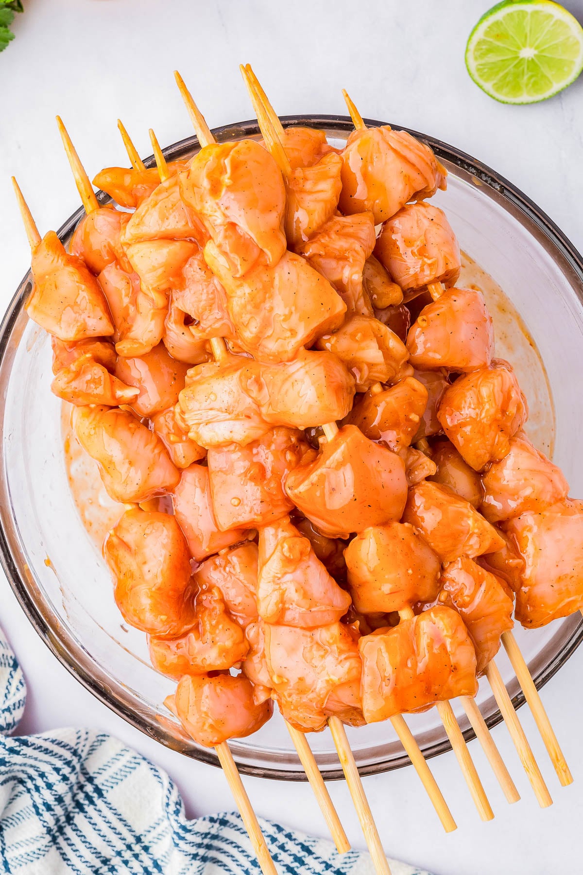 Raw chicken pieces marinated in sauce and skewered on wooden sticks, arranged in a glass bowl on a white surface.