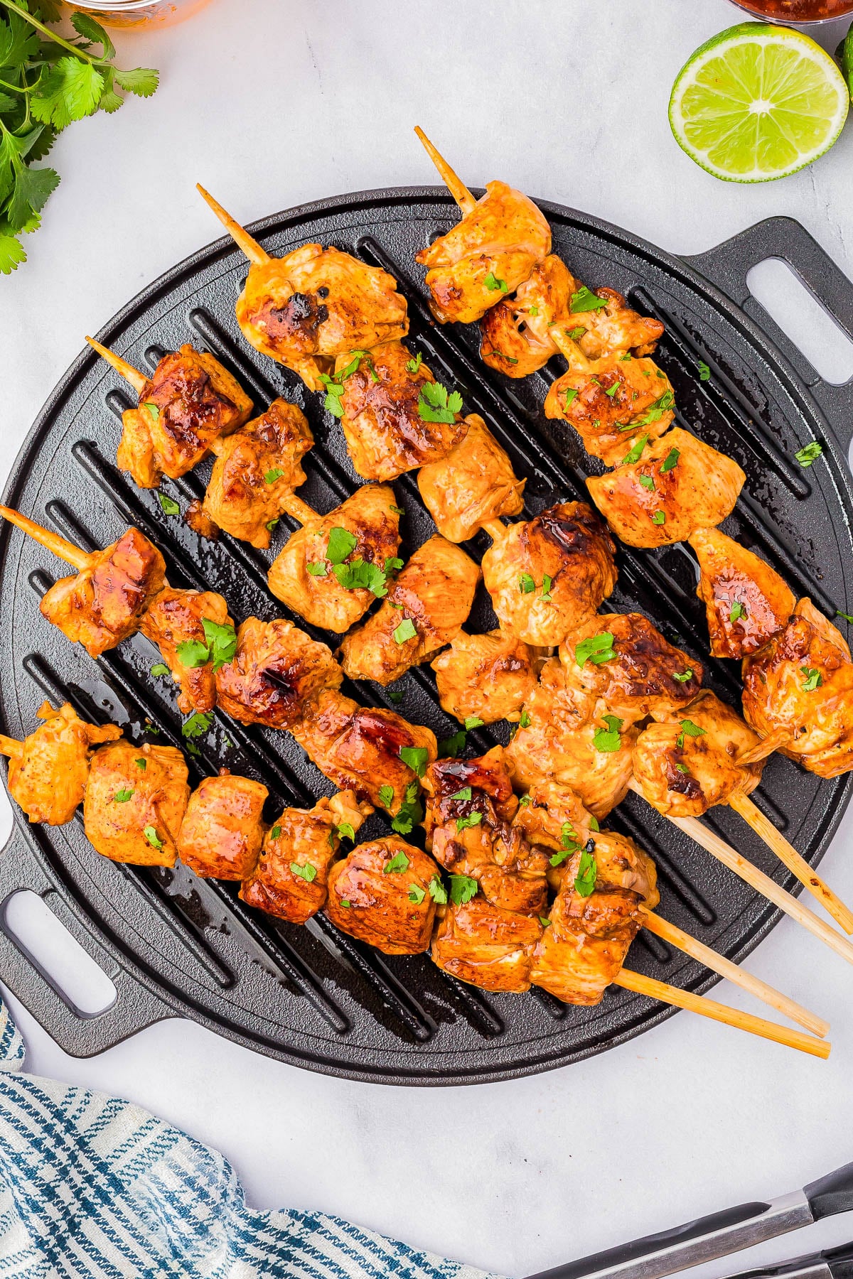 Grilled chicken skewers arranged on a round grill pan, garnished with chopped herbs, with a sliced lime nearby.