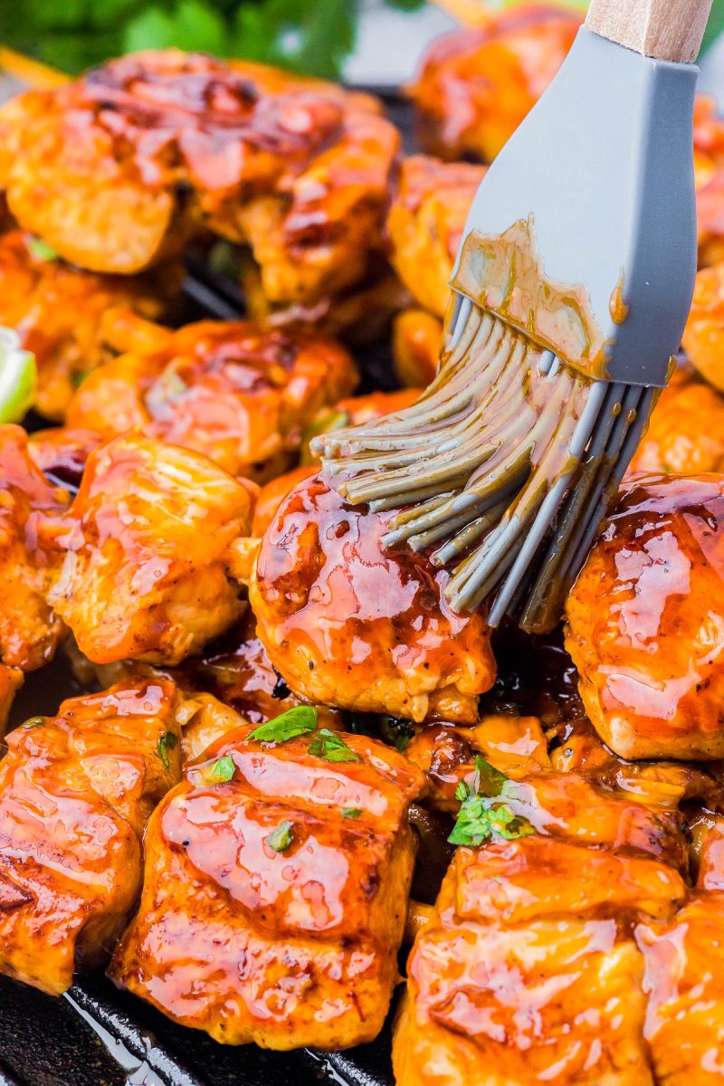 A silicone brush coats grilled chicken pieces with a thick layer of barbecue sauce.