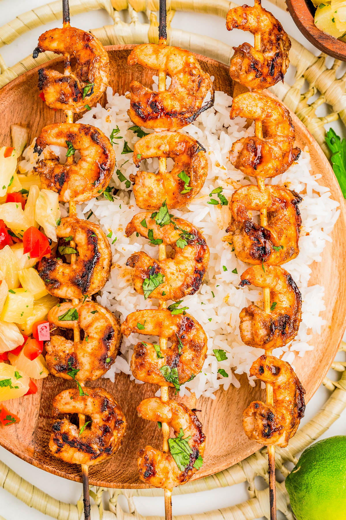 Grilled shrimp skewers served over white rice on a wooden plate, garnished with herbs, with a side of pineapple salsa.