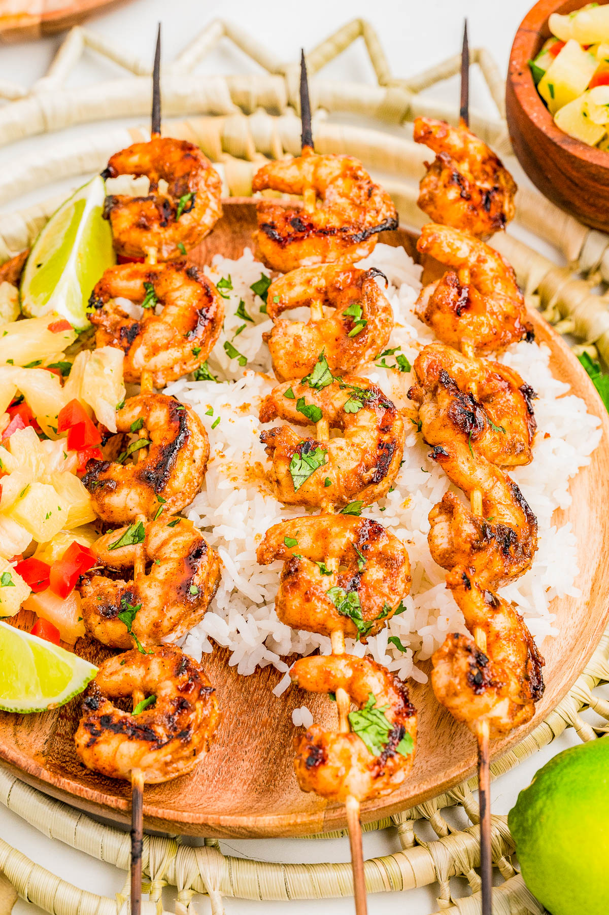 Three skewers of grilled shrimp are served over a bed of white rice, garnished with chopped herbs, lime wedges, and a small side of pineapple salsa.