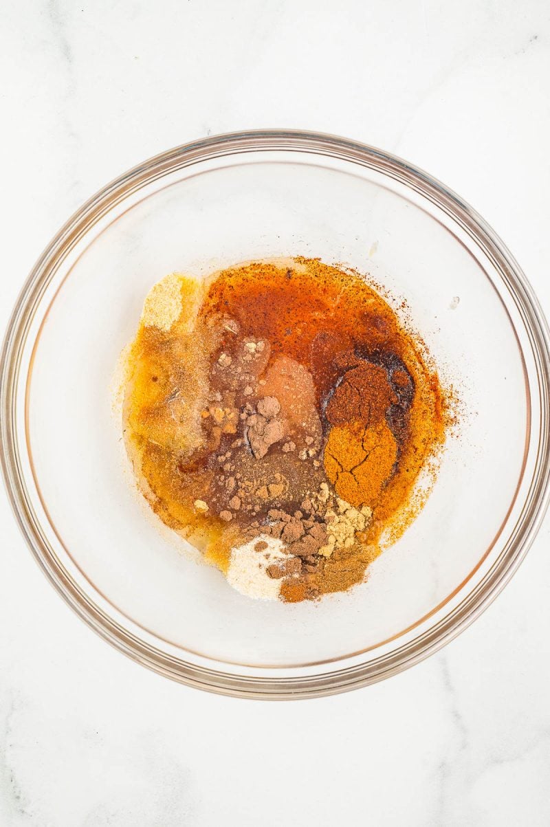 A glass bowl containing a mixture of various ground spices and brown liquid on a white surface.