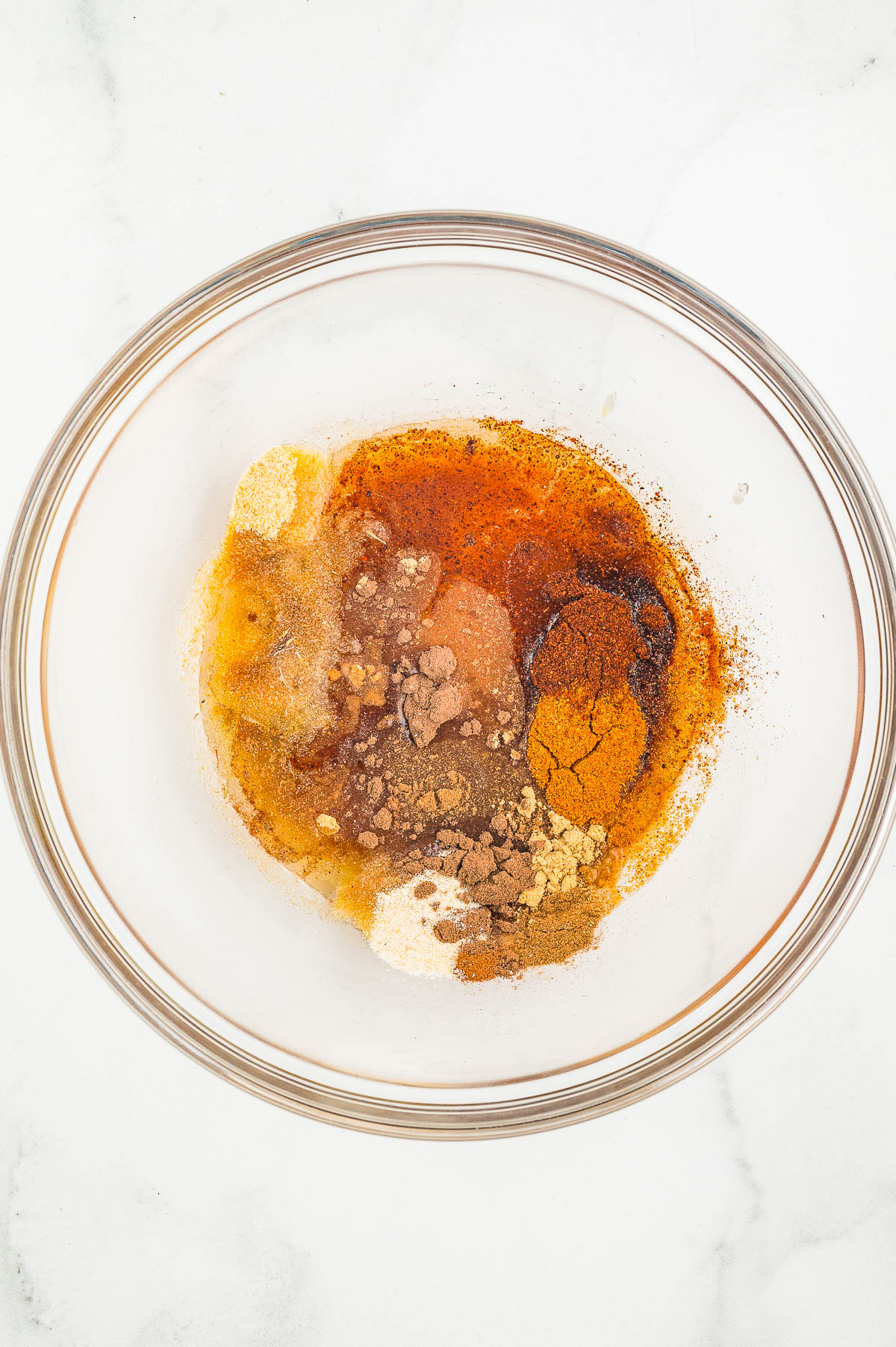 A glass bowl containing a mixture of various ground spices and brown liquid on a white surface.